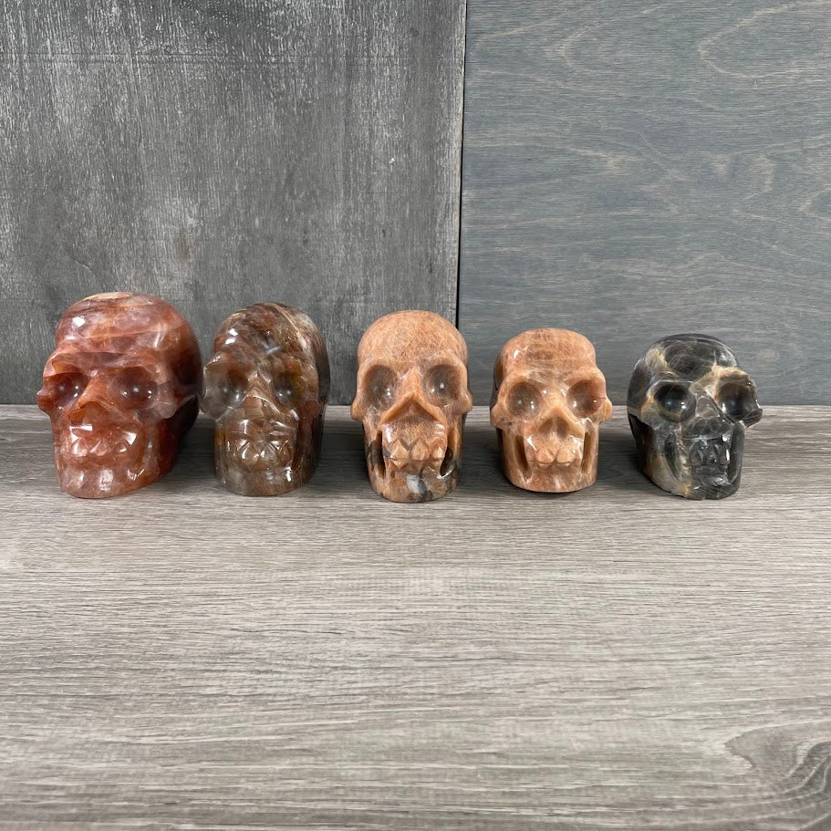 Lot of two peach moonstone skulls, one black moonstone skull, and two hematoid quartz skulls; facing forwards.