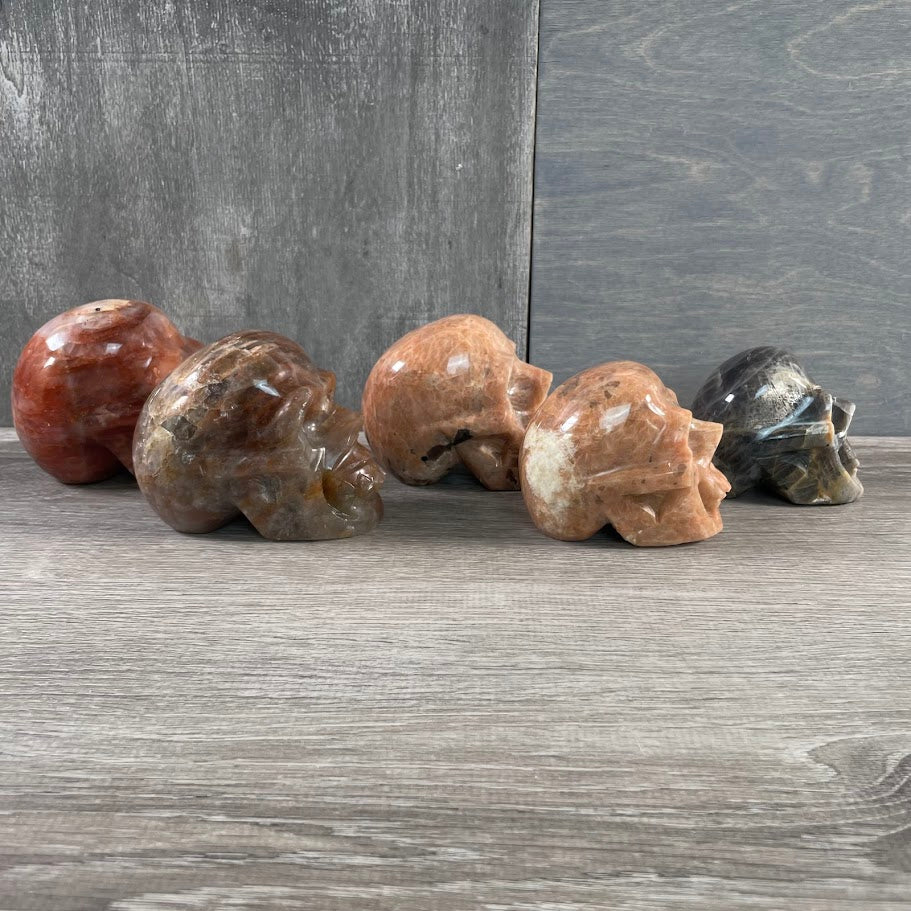 Lot of two peach moonstone skulls, one black moonstone skull, and two hematoid quartz skulls; facing sideways.