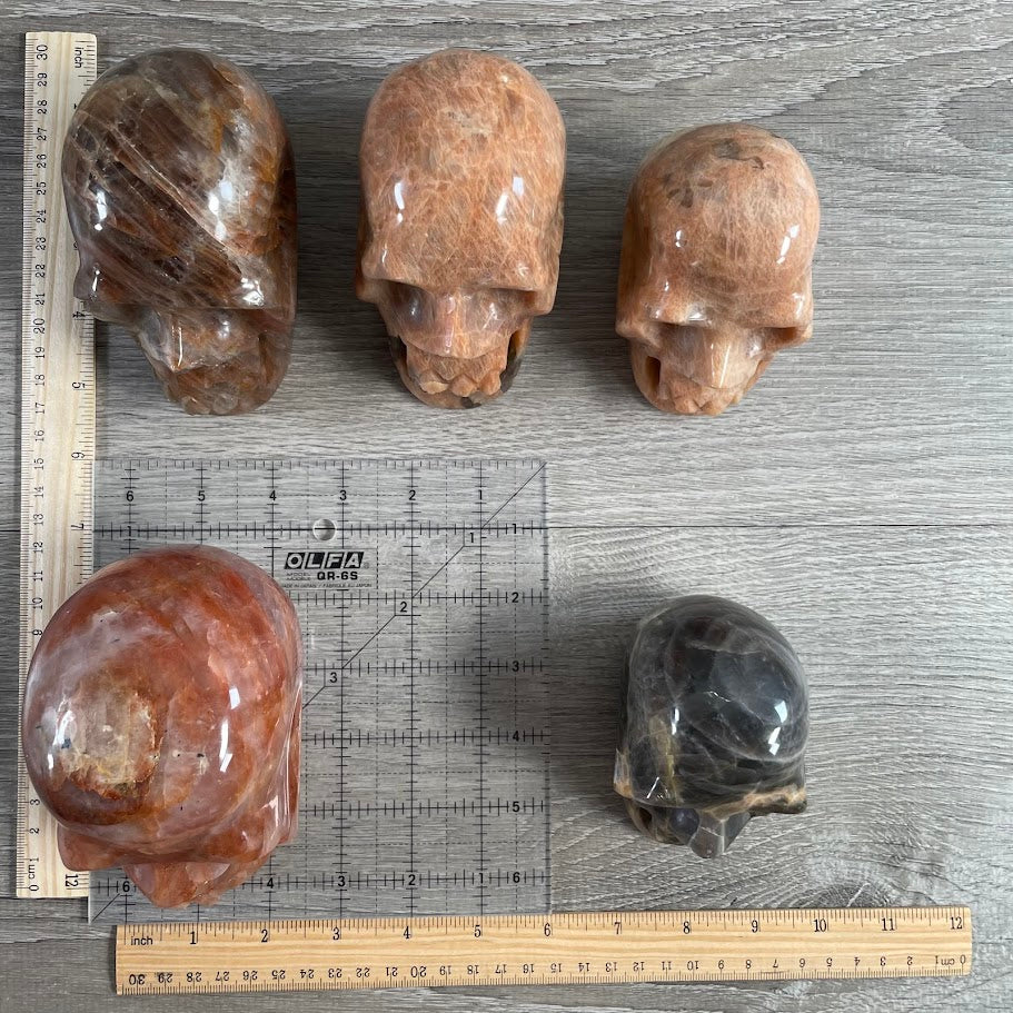 Lot of five crystal skulls: two peach moonstone, one black moonstone, and two hematoid quartz. With grid and rulers for size reference.