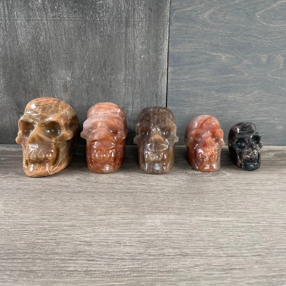 Lot of three hematoid quartz skulls, one black moonstone skull, and one peach moonstone skull; facing forwards.