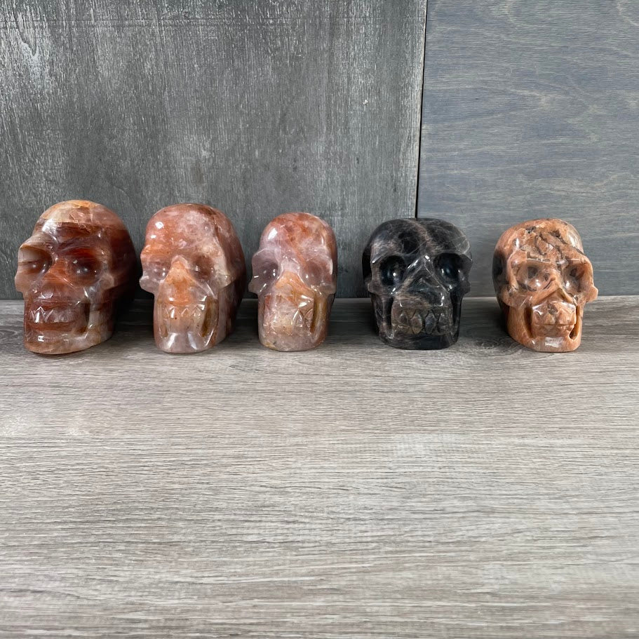 Lot of three hematoid quartz skulls, one peach moonstone skull, and one black moonstone skull; facing forwards.