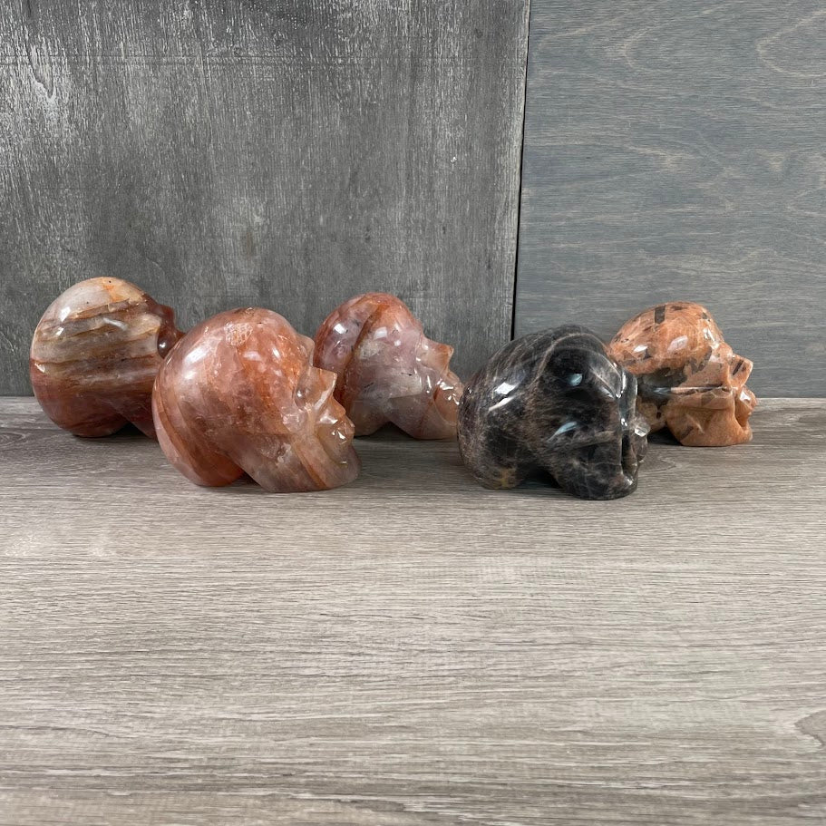 Lot of three hematoid quartz skulls, one peach moonstone skull, and one black moonstone skull; facing sideways.