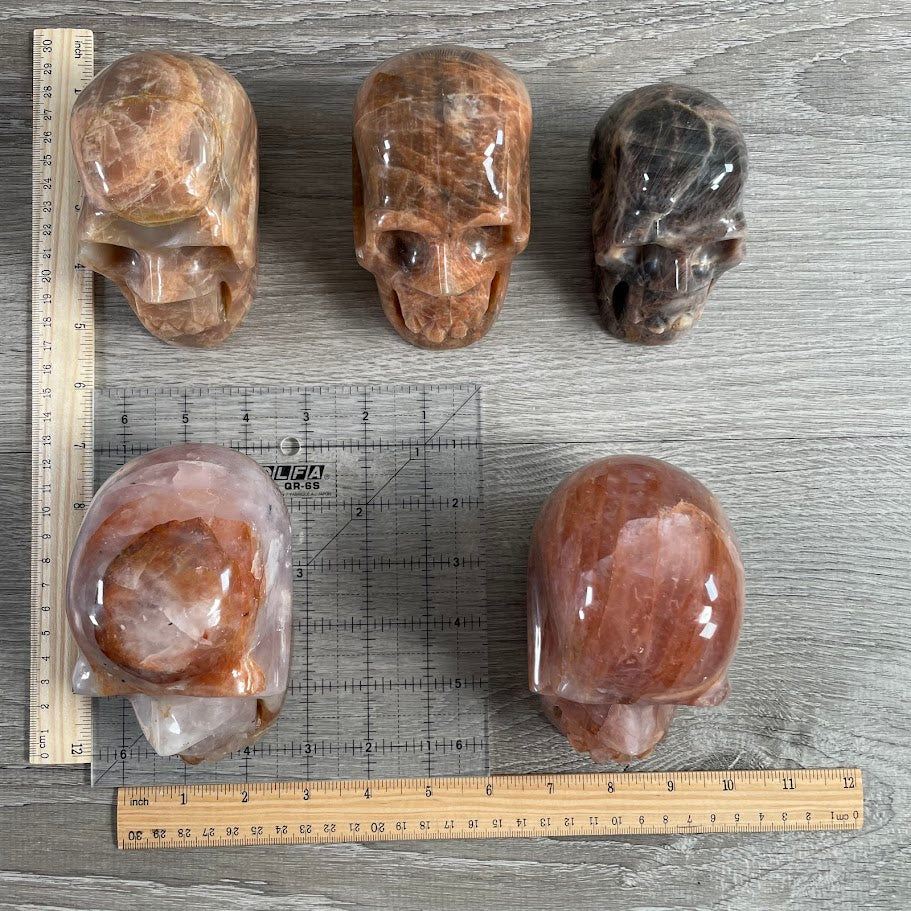 Lot of five crystal skulls: two peach moonstone, one black moonstone, and two hematoid quartz. With grid and rulers for size reference.