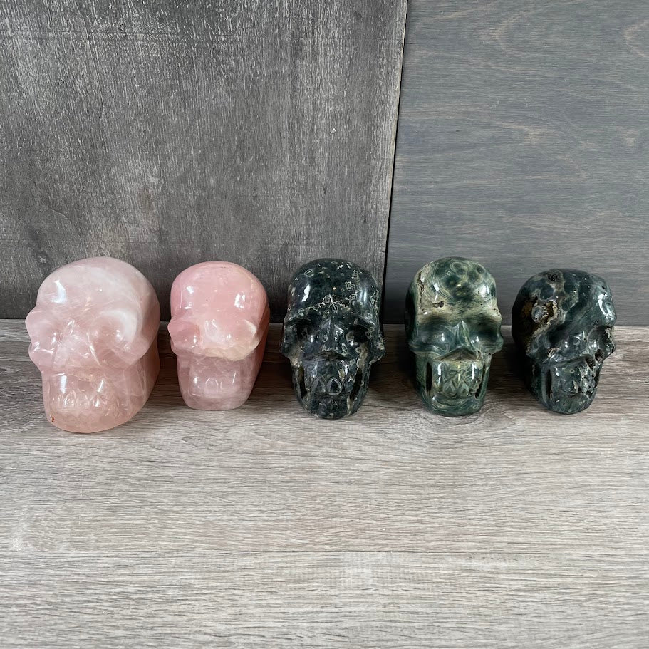 Lot of three Kambaba jasper skulls and two rose quartz skulls; facing forwards.
