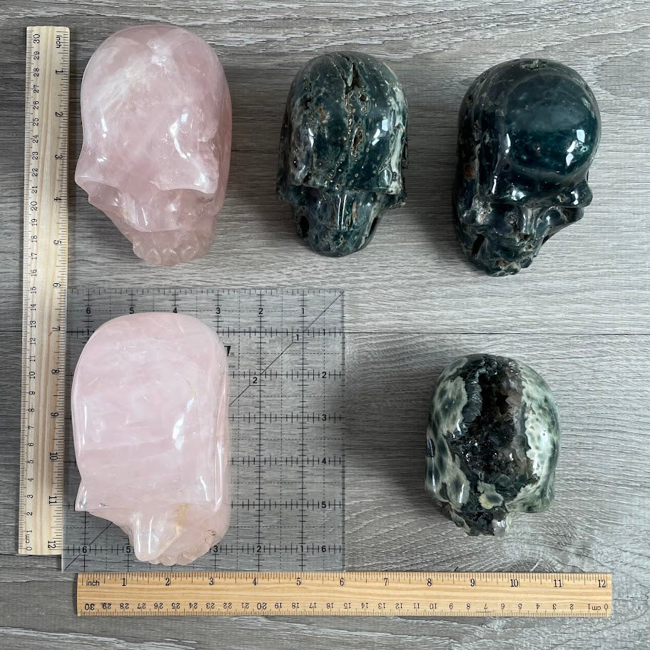 Lot of five crystal skulls: three Kambaba jasper and two rose quartz. With grid and rulers for size reference.