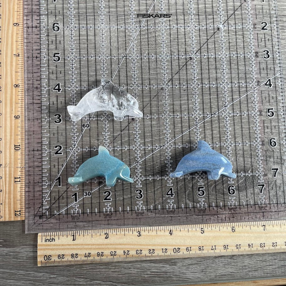 Three brazilian gemstone dolphin figurines, showcasing available variants with a grid and rulers for size reference (each dolphin ~2inch long). 1 blue quartz, 1 quartz, 1 trollite.