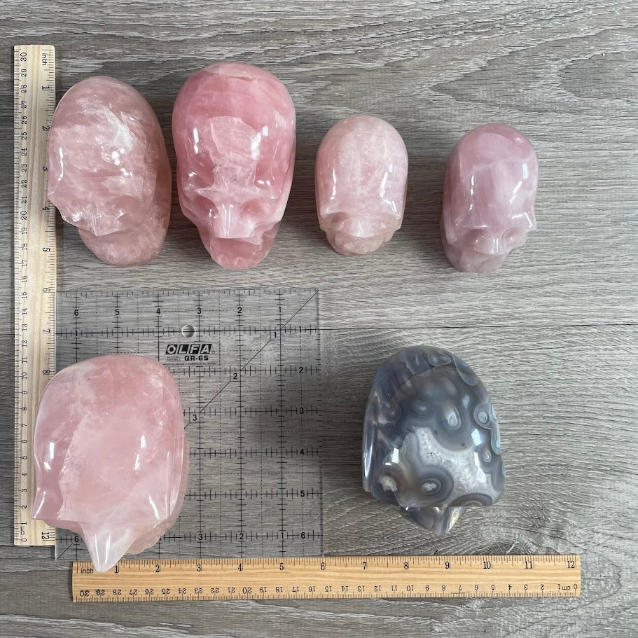 Lot of 6 crystal skulls: five rose quartz skulls and one agate skull. With grid and rulers for size reference. 