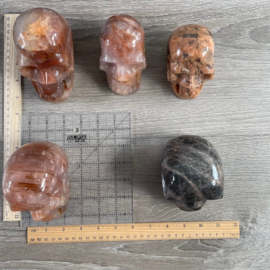 Lot of five crystal skulls: one peach moonstone, one black moonstone, and three hematoid quartz. With grid and rulers for size reference.